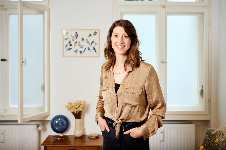 Anna steht in einem Raum vor zwei Fenstern und lächelt in die Kamera